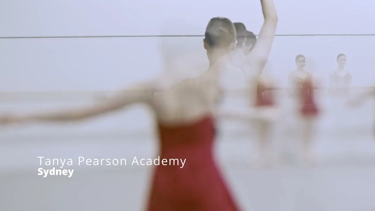 Dancer in a red dress mid-movement in a bright ballet studio, with dancers behind her